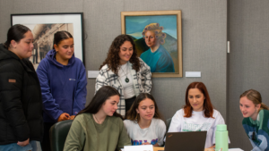 group of young people sitting at a table looking at a computer. Art in background on grey walls.